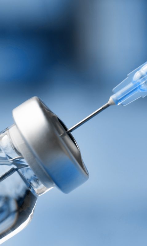 A syringe extracting fluid from a small flask in a pharmaceutical manufacturing setting.